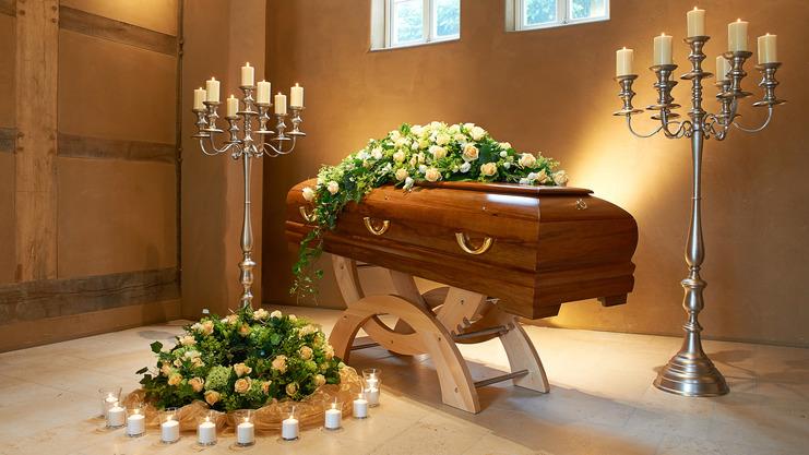 Sarg mit Blumenarrangement in einem Raum, flankiert von Kerzenhaltern und einer Blumendekoration auf dem Boden.
