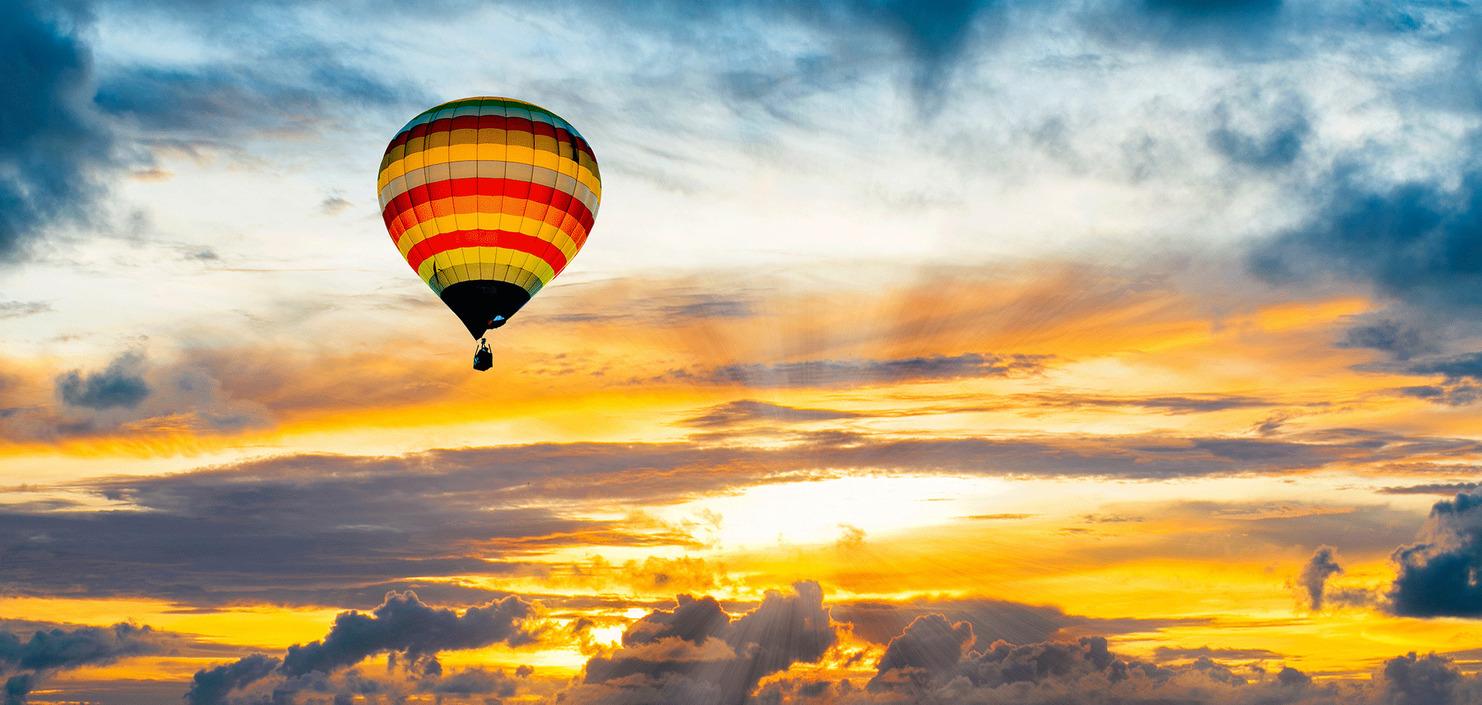 Bunter Heißluftballon schwebt über einem See bei Sonnenuntergang mit dramatischem Wolkenhimmel.