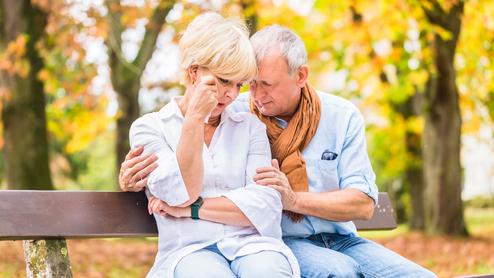 Ein älteres Paar sitzt auf einer Bank im Herbst, mit tröstender Körpersprache und ernsten Gesichtsausdrücken.