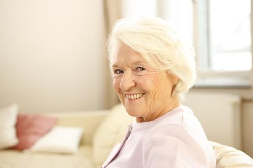 Lächelnde ältere Frau mit grauen Haaren sitzt auf einem Sofa in einem hellen Raum.