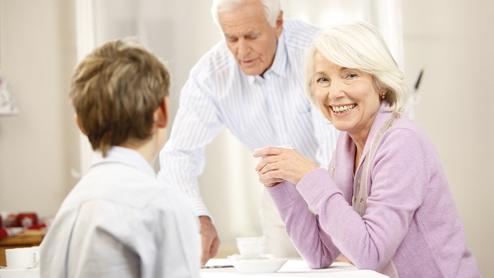 Lächelnde ältere Frau sitzt am Tisch, während ein Mann im Hintergrund Getränke serviert, ein Junge schaut sie an.